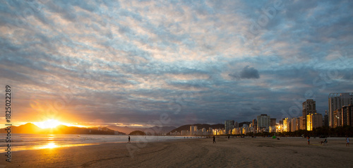 Sunrise over the beach in Santos, Brazil, with dramatic clouds and city buildings reflecting the early light.