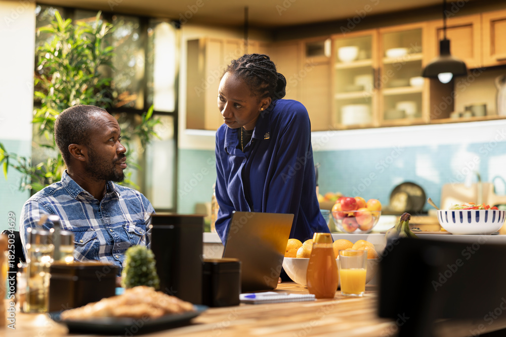 Fototapeta premium African American freelancer sitting at a kitchen table with a laptop, sending emails and explaining business tasks to his girlfriend. Couple enjoys a relaxed home lifestyle with coffee.