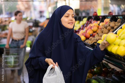 In supermarket,middle-aged woman in traditional Muslim hijab takes and puts ripe apples in small bag