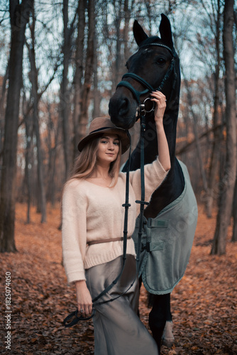 Canvas-taulu Woman stands beside horse in forest during fall season while wearing a hat and s