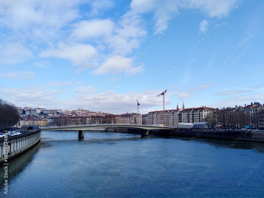 Naklejka premium A bridge crosses the Saône River with the backdrop of Lyon's hill and historic skyline, featuring modern construction with cranes towering over new white buildings.
