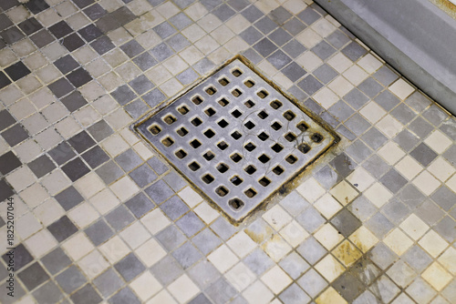 Φωτογραφία A close-up view of a metal square shower drain and grimy checkerboard mosaic floor tiles showing discoloration, mold, and trapped hair