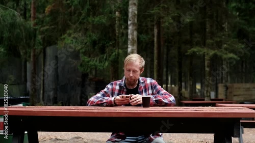 Man with beard checks phone while sitting at wooden table in forest setting