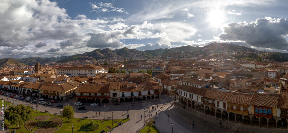 Obraz premium Scenic panorama of the Plaza de Armas in Cusco, Peru, featuring colonial arcades, red tile roofs, and hillside neighborhoods under a dramatic sunny sky with clouds.