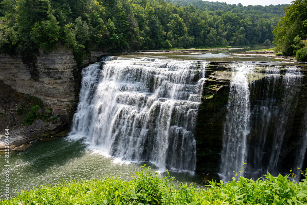 Obraz premium Lush green landscape surrounding a beautiful waterfall cascading down rocky cliffs into a serene lake on a clear sunny day in nature's embrace