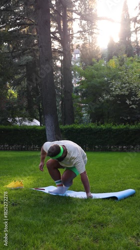 Vertical 4K street video. Fat man with beard, t-shirt, shorts looks asana on mobile phone, raises hand up, stands bend, triangle yoga pose, Trikonasana on blue mat on green grass in city park. Gym day