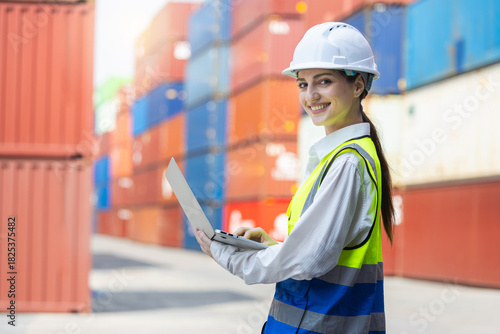 Female engineer using laptop while working at cargo container yard, Smiling industrial worker in safety gear managing logistics at port, Young woman inspecting freight operations with laptop