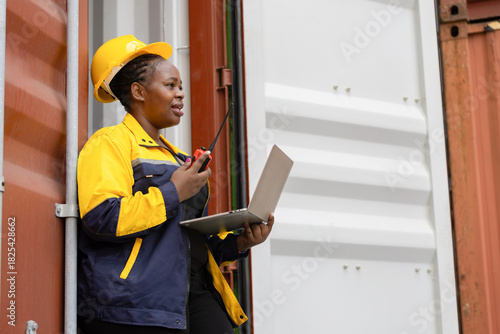 Female industrial worker communicating via walkie talkie while using laptop at container yard, Confident dock worker managing logistics with laptop and radio at shipping terminal