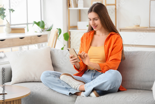 Beautiful young woman with modern mobile phone sitting on sofa at home