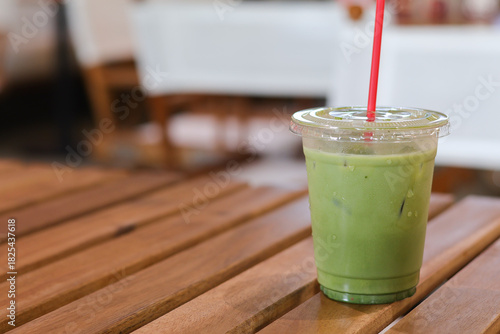 Refreshing iced matcha green tea latte in plastic cup with red straw. This cool beverage sits on wooden table at cafe, perfect drink for relaxing summer day