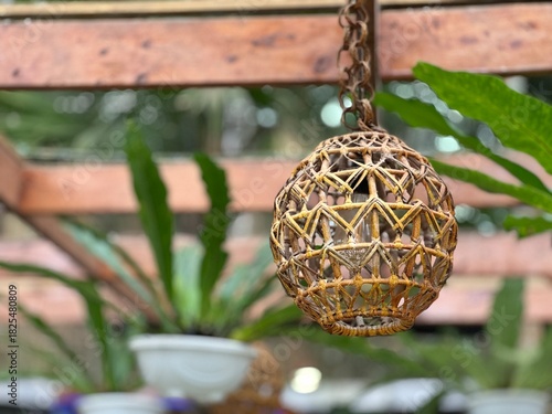 A decorative, handcrafted wicker or rattan ball lantern hangs from a chain, set against a blurred background of wooden beams and lush Bird's Nest Ferns in a warm, natural greenhouse or garden