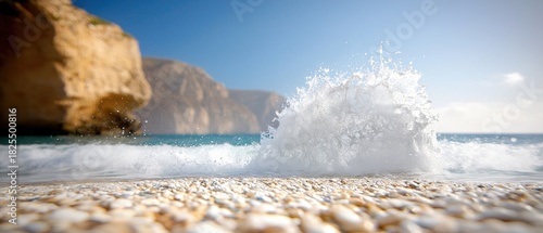 Fototapeta Naklejka Na Ścianę i Meble -  A dynamic close-up shot of a wave crashing onto a pebble beach, with blurred cliffs and a clear blue sky in the background. The water is frothy and white, creat
