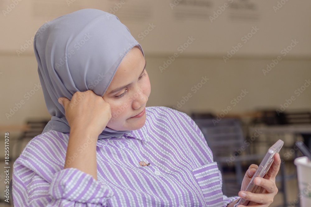 Naklejka premium A young Asian Muslim woman sitting in a coffee shop while waiting for a phone call or message from someone