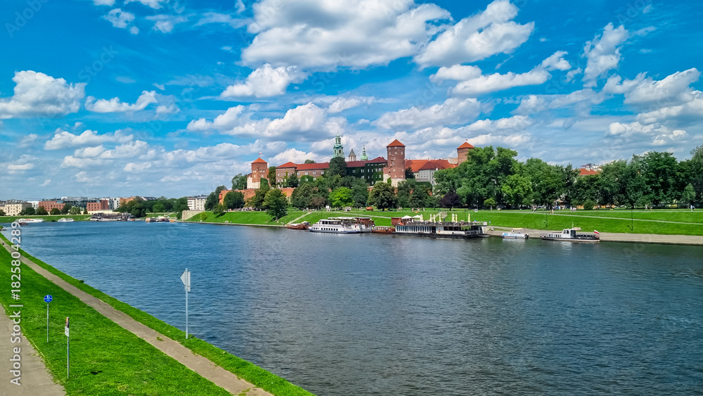 Fototapeta premium Wawel Royal Castle sits proudly on its hill overlooking the Vistula River under a beautiful blue sky with fluffy clouds. Tourist boats are docked at the riverbank, ready for sightseeing tours.