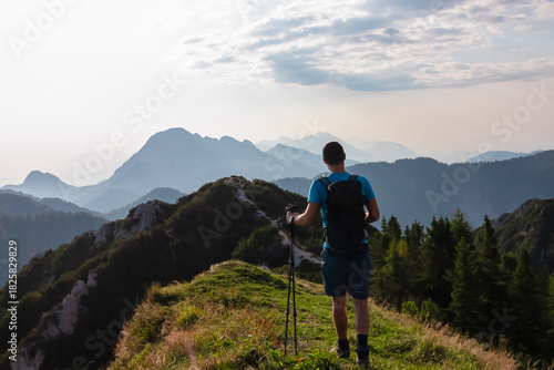 Tableau sur toile A lone hiker with a backpack and trekking poles stands on a grassy summit, admiring the majestic, layered mountain view