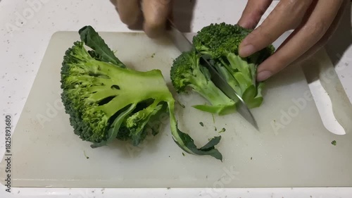 A person is cutting a head of fresh green broccoli into florets on a white cutting board in a bright kitchen setting