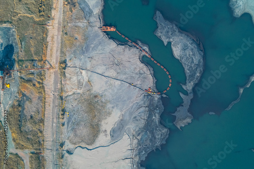 Aerial abstract composition of a mining tailings pond. A dredge pipe snakes across grey sludge and toxic blue water, highlighting severe industrial pollution and technological impact.