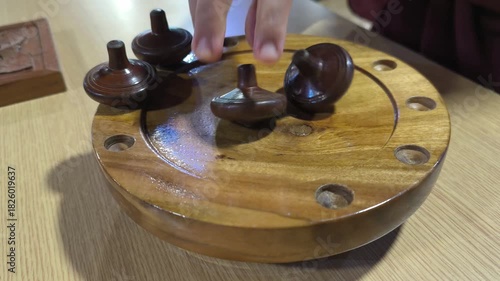 Small wooden traditional Malay spinning tops are spun on a wooden platform that slopes to the center. Multiple tops are competed in this activity, representing a vanishing traditional Malay game.