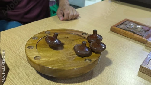 Small wooden traditional Malay spinning tops are spun on a wooden platform that slopes to the center. Multiple tops are competed in this activity, representing a vanishing traditional Malay game.