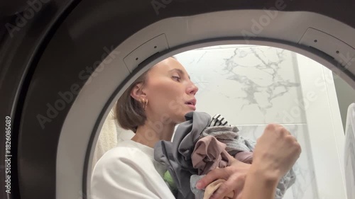 Young woman unloading clothes from tumble dryer – inside view through washing machine drum in modern bathroom. Household chores, home routine, laundry process