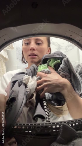 Young woman unloading clothes from tumble dryer – inside view through washing machine drum in modern bathroom. Household chores, home routine, laundry process
