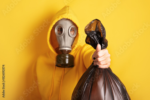 Individual in yellow hoodie and gas mask holding a black garbage bag against a vibrant yellow background. Concept of pollution or waste management.