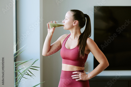 Fotografía Young adult Caucasian woman standing near window drinking green smoothie with ey