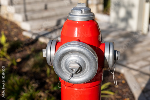 Red outdoor fire hydrant with metal caps in a residential area, photographed in sunlight. Safety equipment for firefighting and emergency water access.