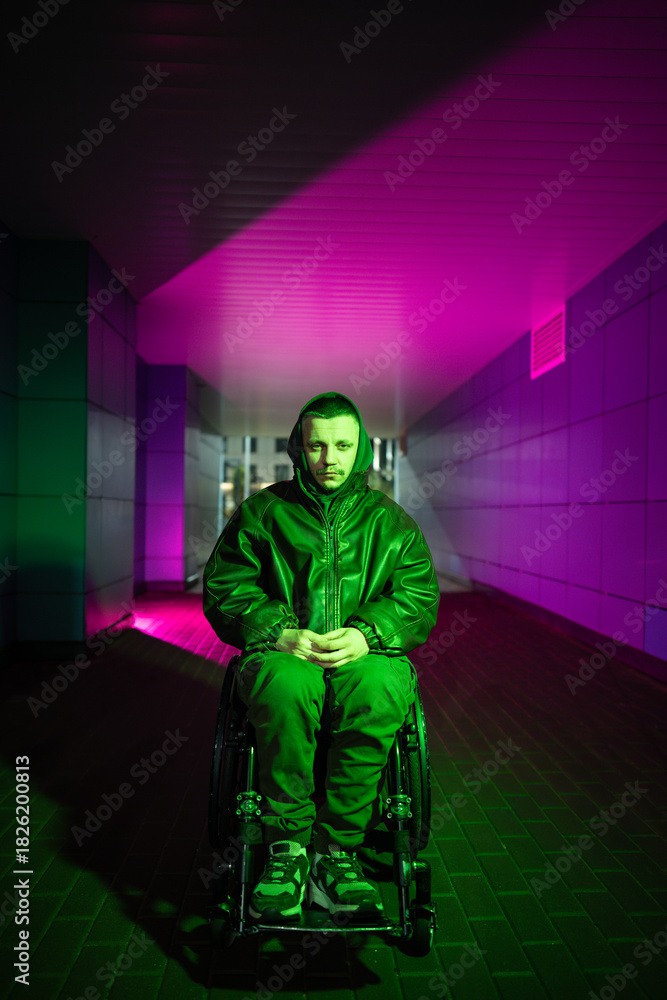 Obraz premium Pensive young disabled man in wheelchair walking alone, sitting in color neon rgb light looking at camera with tired sad expression. Male with disability showing signs of heavy mental state and apathy