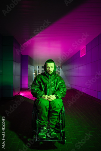Pensive young disabled man in wheelchair walking alone, sitting in color neon rgb light looking at camera with tired sad expression. Male with disability showing signs of heavy mental state and apathy