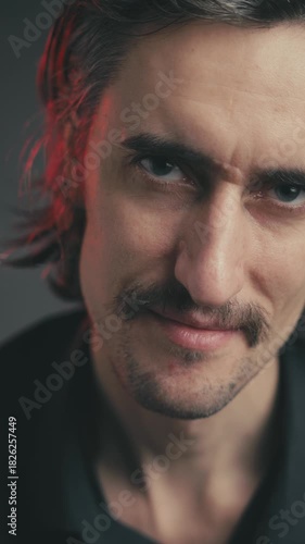 Angry look, sly, cunning, serious, dangerous young attractive European man with a mustache and long hair conveys deep emotions through subtle facial expressions in moody lighting.