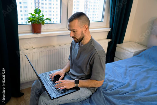 Quadro su tela A man is lying down on his bed in a bedroom, focused on typing on his laptop