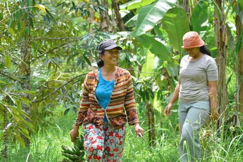 A mother carries bananas and a sickle, smiling brightly while her teenage daughter follows behind in the lush agroforestry field.