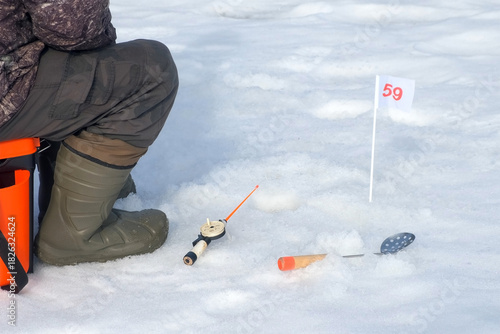 Ice fisherman sitting patiently with rod and flag indicator on fishing competition. Man catching fish on frozen river with ice and snow. Outdoor winter activity hobby leisure recreation kind of sport.