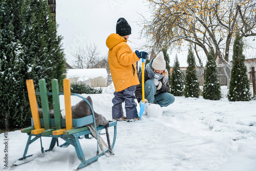 Mother and child playing in a snowy backyard, building a snowman with sled and shovel; cozy winter games, eco-friendly screen-free family time, candid seasonal lifestyle