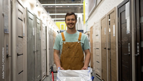 Happy construction worker wearing overalls pushes a cart through a hardware store aisle, smiling at the camera while shopping for doors and building supplies