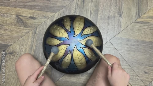 Close up top view of child sitting on wooden floor playing metal tongue drum with mallets creating soft meditative sound. Suitable for mindfulness, kids music learning, calm atmosphere, Concept of