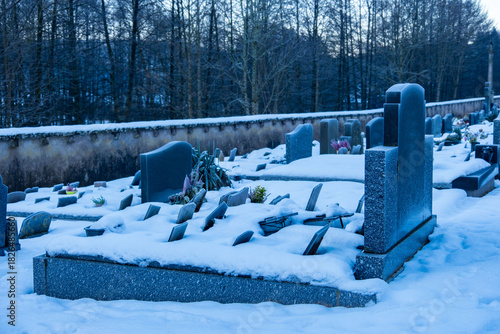 Snowy cemetery scene with old stone wall, cross, and metal gate, set against backdrop bare trees and cold blue sky, evoking peace, memory, and quiet beauty of winter