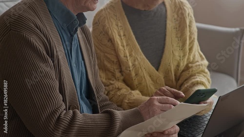 Tilt down shot of senior Caucasian couple analyzing household paperwork while using smartphone and laptop, sitting on sofa in cozy living room at daytime