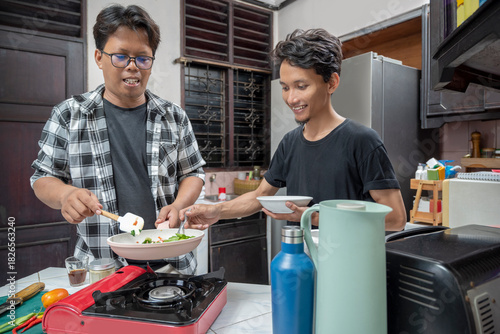 Two Indonesian southeast asian men are cooking together in a kitchen. Cooking in a frying pan on a gas stove, while holding a plate tasting the dish with a spoon. Cooking activity