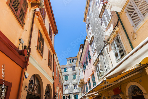 Fototapeta Naklejka Na Ścianę i Meble -  Sun-drenched narrow street in Corfu, Greece, framed by pastel buildings, wooden shutters, and clear blue sky.