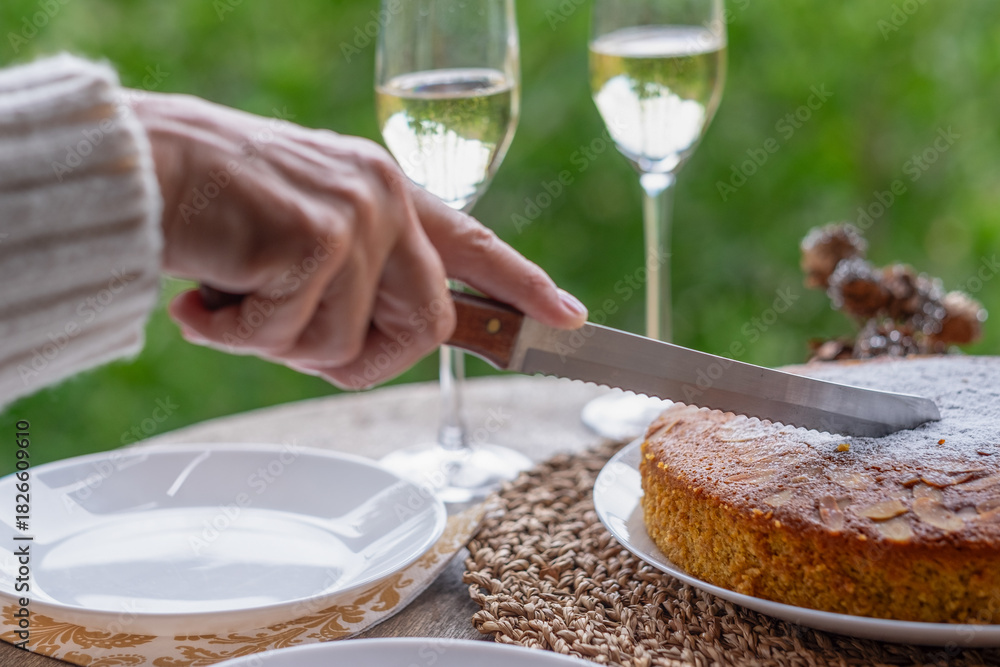 Obraz premium Rustic table on balcony and woman's hand cutting a slice of homemade carrot cake with almonds. A glass of wine to celebrate party or birthday in winter season