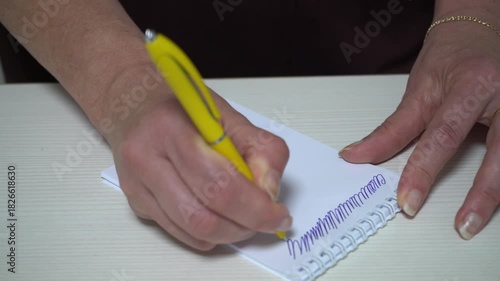 A person practices handwriting using a yellow pen on a blank notebook. The setting is a simple desk, showcasing the focus on writing skills.