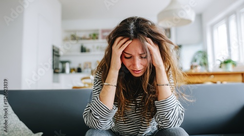 Stressed woman holding head with both hands sitting on couch in bright room, expressing frustration, anxiety, tension, overwhelmed emotions, mental strain, fatigue, and personal stress concept