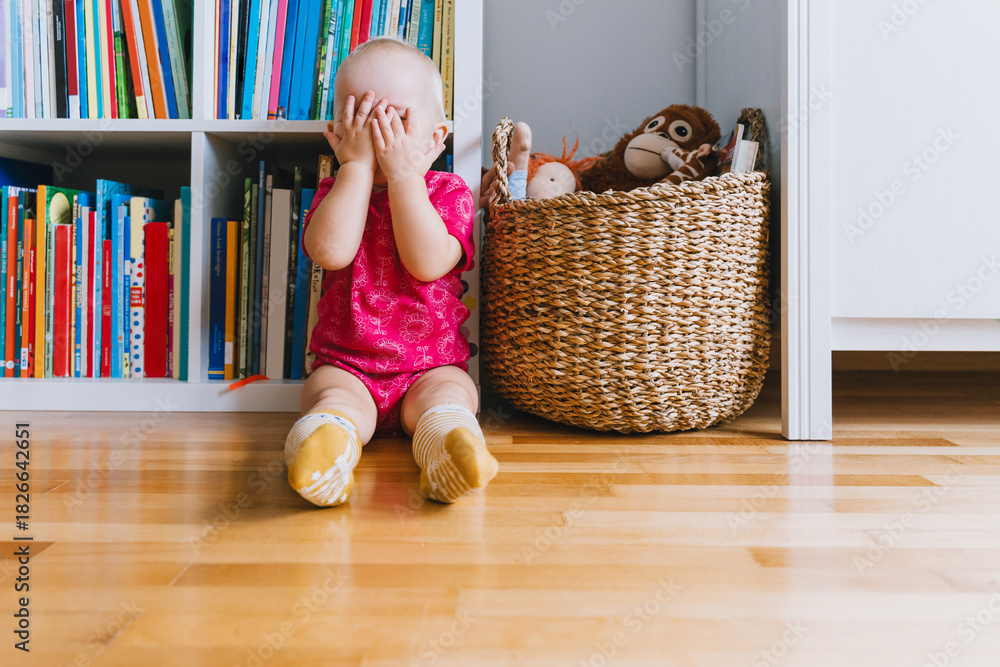 Ταπετσαρία Little cute girl playing peekaboo sitting on floor in front of bookshelf and basket of toys in baby room or daycare