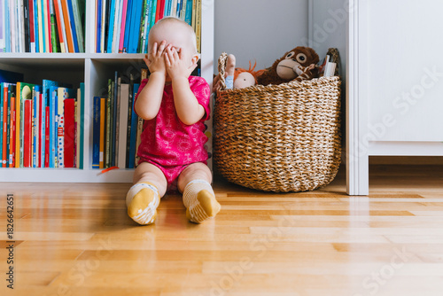 Little cute girl playing peekaboo sitting on floor in front of bookshelf and basket of toys in baby room or daycare. Toddler playing indoors. Family with one child at home. Happy childhood.