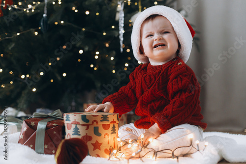 Cute baby in santa hat crying at present boxes with garland lights under modern decorated christmas tree. Sad overwhelmed baby. Holiday time