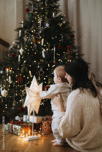 Stylish mother and baby son in knitted sweaters sitting together against decorated christmas tree with lights and presents. Atmospheric holiday time, Merry Christmas! Baby's first christmas