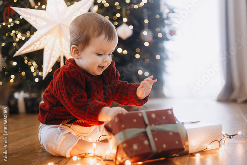 Merry Christmas and Happy Holidays! Cute baby in knitted sweater sitting with stylish gift box and lights at decorated christmas tree and star. Atmospheric magical moment