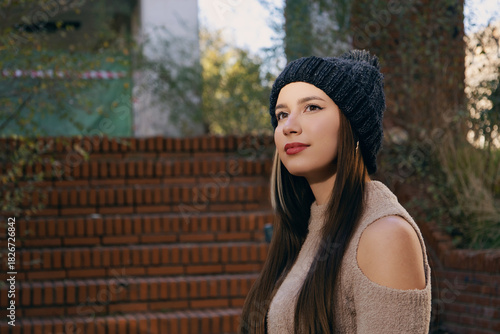 Mujer joven con gorro y bufanda de invierno posando casualmente en parque urbano soleado.